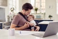 Transgender Father Working From Home On Laptop Looking After Baby Son In Sling Royalty Free Stock Photo