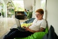 Transgender Gen Z individual smiles while working on laptop seated on green bean bag in airy office. Inclusive, casual