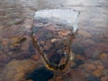 Translucent angular clean piece of ice in running water with colorful stones underneath in winter, rigidity, stillness and