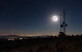 Transmission masts on a high mountain under a full moon with the illuminated coastline.