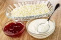 Transparent bowl with cottage cheese, bowl with raspberry jam, spoon in bowl with sour cream on wooden table Royalty Free Stock Photo