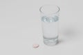 A transparent glass filled with drinking water. Next to the glass of water is a round vitamin powder. The background is white