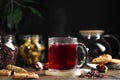 Transparent mug with hot red tea on a dark background, near cookies and tea leaves