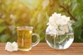 Transparent mug of tea and a vase with jasmine on a wooden table, greens on the background Royalty Free Stock Photo