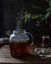 Transparent teapot with herbal tea on a wooden table. Steam over the kettle. Flowering branches in the background.