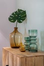 Transparent yellow and green vases for flowers and big bottle with monstera leaf on wooden cupboard. Decoration of