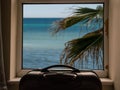 Travel case by a dirty window with view on blue water and palm tree, selective focus, Cheap budget holiday concept