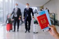 Travel concept: hand holding passport, mask, boarding pass in airport. Two pilots walking with luggage in background Royalty Free Stock Photo