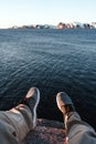 Travel and lifestyle concept. Brave traveler hipster sits on cliff in front of ocean and mountain. POV view on legs and shoes on b