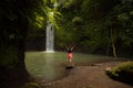 Travel lifestyle. Young traveler woman wearing pink dress at waterfall in tropical forest. Excited woman raising arms in front of Royalty Free Stock Photo