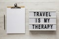 `Travel is my therapy` words on a modern board, clipboard with blank sheet of paper on a white wooden surface, top view. Overhea