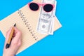 Travel planning, vacation concept. A hand holds a pen over an open blank notebook, dollars, pink sunglasses Royalty Free Stock Photo
