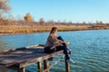 Traveler with backpack relaxing by autumn river at sunset. Young woman sitting on pier admiring landscape Royalty Free Stock Photo