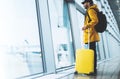 Traveler in bright jacket with yellow suitcase backpack at airport on background large window blue sky, passenger waiting flight Royalty Free Stock Photo