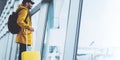 Traveler in bright jacket with yellow suitcase backpack at airport on background large window blue sky, passenger waiting flight Royalty Free Stock Photo