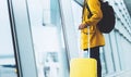 Traveler in bright jacket with yellow suitcase backpack at airport on background large window blue sky, passenger waiting flight Royalty Free Stock Photo