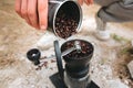 Traveler filling grinder with fresh coffee beans on cliff at lake, preparing for brewing alternative coffee at camping. Making hot