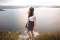 Traveler girl in hat and with windy hair standing on top of rock mountain, enjoying beautiful sunset view on river. Young woman in Royalty Free Stock Photo