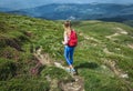 Traveler girl with red backpack is walking along green path in mountains. Summer or spring flowers rue Royalty Free Stock Photo