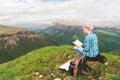 A traveler girl sits in the mountains on the grass and reads a book on the background of epic mountains. The concept of