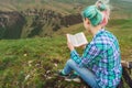 A traveler girl sits in the mountains on the grass and reads a book on the background of epic mountains. The concept of