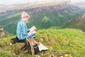 A traveler girl sits in the mountains on the grass and reads a book on the background of epic mountains. The concept of