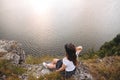 Traveler hipster girl in hat and windy hair sitting on top of rock mountain with wildflowers, looking at beautiful sunset view on Royalty Free Stock Photo
