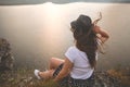 Traveler hipster girl in hat and windy hair sitting on top of rock mountain with wildflowers, looking at beautiful sunset view on Royalty Free Stock Photo