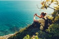 Traveler man sits on shore and takes pictures of sea on cell phone camera during sunset