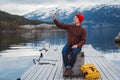Traveler man taking self-portrait a photo with a smartphone. Tourist in a yellow backpack sitting on wooden pier a Royalty Free Stock Photo