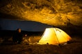 Traveler near the glowing camping tent in the night grotto under a starry sky