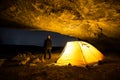 Traveler near the glowing camping tent in the night grotto under a starry sky