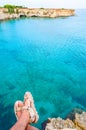 Sitting on the edge of a cliff with dangling legs in travel sandals looking on crystal clear water on Torre Sant Andrea beach with