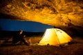 Traveler with smartphone near the glowing camping tent in the night grotto under a starry sky