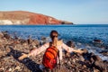 Traveler woman with backpack feels happy raising arms on Red beach on Santorini island, Greece enjoying landscape Royalty Free Stock Photo