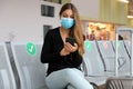 Traveler woman with surgical mask using smart phone and sitting respecting social distancing at the airport lounge