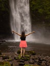 Traveler woman wearing pink dress at waterfall. Excited woman raising arms in front of waterfall. Travel lifestyle. Energy of Royalty Free Stock Photo