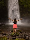 Traveler woman wearing pink dress at waterfall. Excited woman raising arms in front of waterfall. Travel lifestyle. Energy of Royalty Free Stock Photo