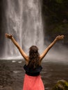 Traveler woman wearing pink dress at waterfall. Excited woman raising arms in front of waterfall. Travel lifestyle. View from back Royalty Free Stock Photo