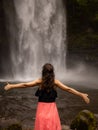 Traveler woman wearing pink dress at waterfall. Excited woman raising arms in front of waterfall. Travel lifestyle. View from back Royalty Free Stock Photo