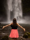 Traveler woman wearing pink dress at waterfall. Excited woman raising arms in front of waterfall. Travel lifestyle. View from back Royalty Free Stock Photo