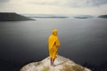 Traveler in yellow raincoat standing on cliff and looking at lake in rainy windy day. Wanderlust and travel concept. Hipster man Royalty Free Stock Photo