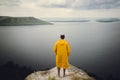 Traveler in yellow raincoat standing on cliff and looking at lake in rainy windy day. Wanderlust and travel concept. Hipster man Royalty Free Stock Photo