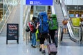 Travelers with backpacks and suitcase go to the escalator in lobby of airport, people queue up at check-in counters for flight,