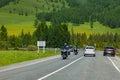 Travelers on motorcycles and cars drive in a sharp turn on an asphalt road towards cars near mountains with green grass and trees