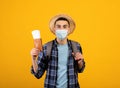 Traveling during covid. Young tourist with backpack wearing face mask, holding tickets and passport, orange background