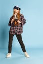 Traveller. Young beautiful girl in checkered shirt, cap and sunglasses posing with photo camera over blue studio Royalty Free Stock Photo