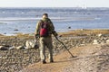 A treasure hunter with a metal detector walks along the sandy deserted beach in search of lost coins