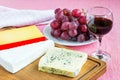 Tree kinds of cheese on brown wooden cutting board, glass of red wine and sweet red grapes on white plate. Cheeses covered with Royalty Free Stock Photo