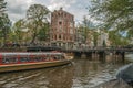Tree-lined canal with bridge, touristic boat, buildings and cloudy sky in Amsterdam.